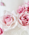 A close-up of a white decorated cake or tower covered with large pale pink roses and small buds, set against a soft light gray background.