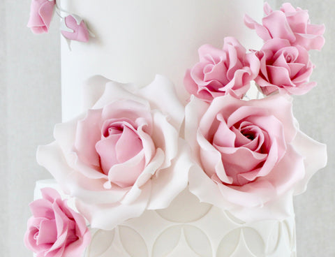 A close-up of a white decorated cake or tower covered with large pale pink roses and small buds, set against a soft light gray background.